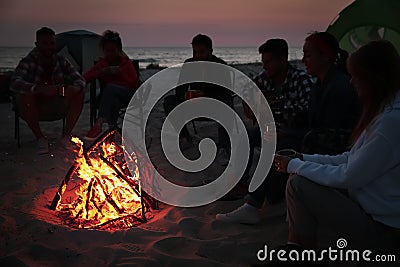 Group of friends gathering around bonfire on beach in evening. Camping season Stock Photo