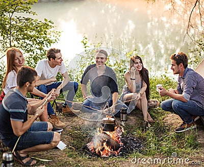 Group of friends are sitting around camp fire and preparing sausages. Stock Photo