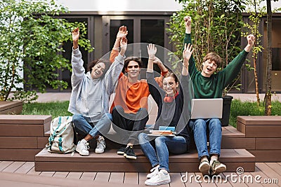 Group of joyful students sitting and happily looking in camera while raising their hands up in courtyard of university Stock Photo
