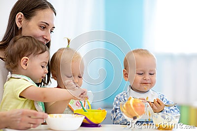 Group of nursery babies eating healthy food with babysitter help. Lunch break in creche. Time to eat in daycare Stock Photo