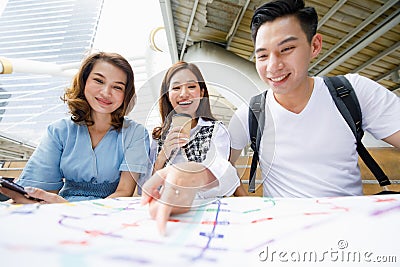 Group portrait of three adult Asian tourists sitting on the stair and holding paper map with a woman holding disposable coffee cup Stock Photo