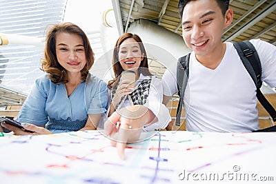 Group portrait of three adult Asian tourists sitting on the stair and holding paper map with a woman holding disposable coffee cup Stock Photo