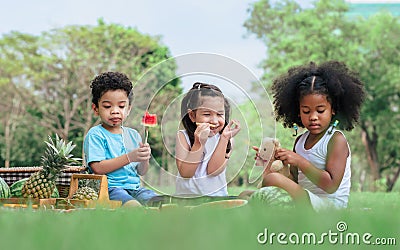 Group of three mixed race African and caucasian little cute kids sitting, playing in outdoor green park for picnic, eating fruit, Stock Photo