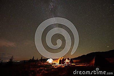 Group of tourists with guitar by burning bonfire under dark starry sky with Milky Way constellation. Stock Photo