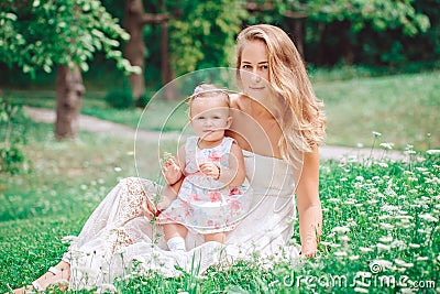 Group of two people, white Caucasian mother and baby girl child in white dress sitting playing in green summer park forest outside Stock Photo
