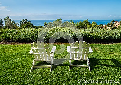 Group of two white Adirondack chairs on green lawn facing a hedge and ocean Stock Photo