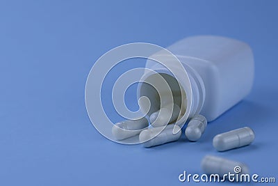 A group of white capsules of medicine spilled out of a white jar on a blue background. Stock Photo