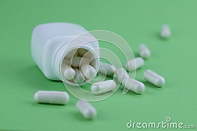 A group of white capsules of medicine spilled out of a white jar on a green background. Stock Photo