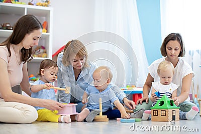 Group of workers with babies in nursery or kindergarten. Moms playing with kids in creche Stock Photo