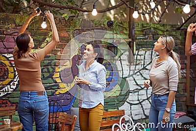 Group of young adult caucasian women setting up hanging bulbs on branches in the garden, preparing for party Stock Photo