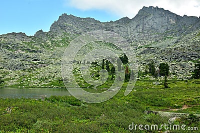 A group of young cedars on the rocky shore of a wide lake surrounded by high rocky mountains with pointed peaks on a sunny summer Stock Photo