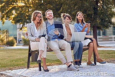 Group of young people laughing together in park, sitting on bench, holding their cell phones, laptop, tablet. sharing good news Stock Photo