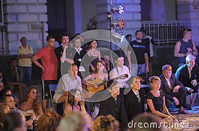 Group of young people sitting on karemats outdoor watching the play of the street theatre actors, girl playing guitar in Editorial Stock Photo