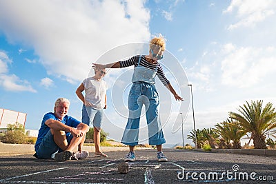 Grouyp of threee people of different ages playing together at the hopscotch on the asphalt of the street - seniors and adult woman Stock Photo