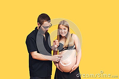 the guy draws with cream emoji on the belly of his pregnant wife on a yellow background Stock Photo