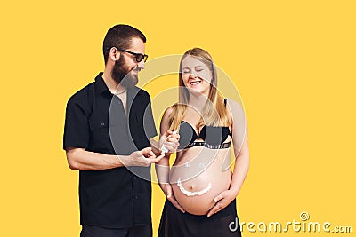 the guy draws with cream emoji on the belly of his pregnant wife on a yellow background Stock Photo