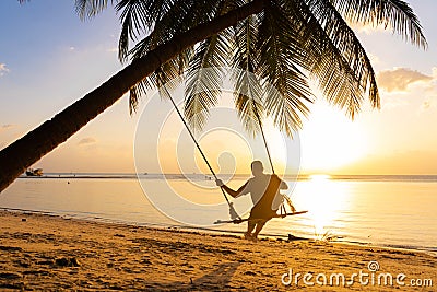 The guy enjoys the sunset riding on a swing on the ptropical beach. Silhouettes of a guy on a swing hanging on a palm tree, Stock Photo