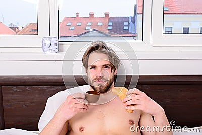 Guy having coffee and holds croissant while lay bed in bedroom or hotel room. Man eats croissant and drinking coffee Stock Photo