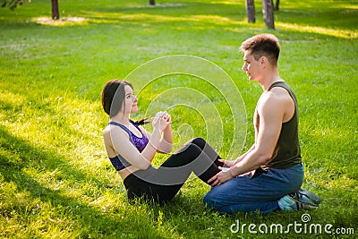 guy helps the girl to do the press exercise, sitting on the lawn in the park Stock Photo