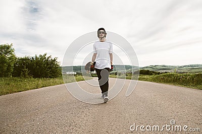 A guy with a longboard in his hands in a helmet and sunglasses is walking along an asphalt suburban road Stock Photo