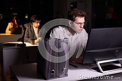 Guy realising amount of work in office at night, sitting at desk working on computer Stock Photo