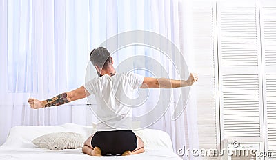 Guy stretching arms, full of energy in morning, rear view. Man in shirt sits on bed, white curtains on background. Macho Stock Photo