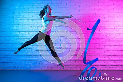 Gymnast woman stretching in front of brick wall in neon lights. Flexible muscular woman doing spirals with ribbon. Stock Photo