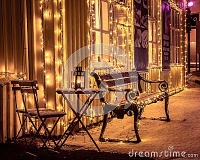 A wooden bench, a table with decorative candle lantern covered with snow and illuminated with Christmas lights. Editorial Stock Photo