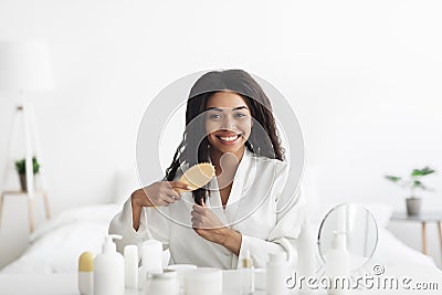 Daily hair care at home during covid-19 lockdown. Happy african american lady combing her curly hair in bedroom Stock Photo