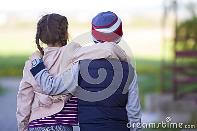 Half-length back view of two children, girl with long dark braids and boy hugging each other over shoulders on blurred outdoors Stock Photo