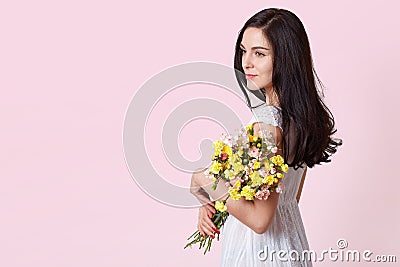 Half length portrait of charming brunette woman in white sundress holding bouquet of flowers over pink background, looking Stock Photo