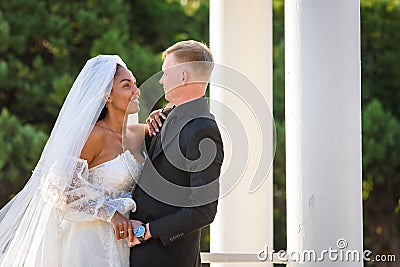 Half-length portrait of mixed-racial newlyweds on a walk against the background of columns and greenery Stock Photo