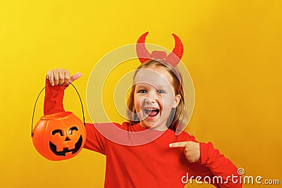 Halloween celebration. Little girl in a devil costume on a yellow background. The child holds a pumpkin jack lantern Stock Photo