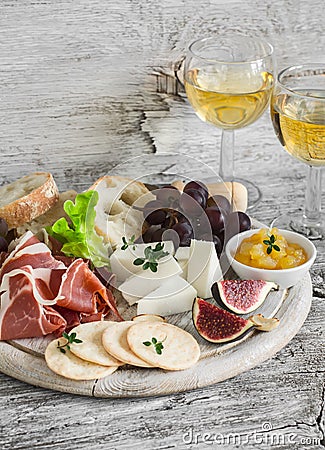 Ham, cheese, grapes, figs, nuts, bread ciabatta, cracker, jam on white wooden board and two glasses of white wine on bright wooden Stock Photo