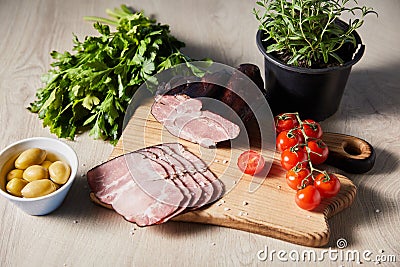 Ham on cutting board with greens, cherry tomatoes, olives on wooden table Stock Photo