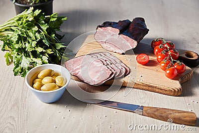 Ham on cutting board with knife, parsley, cherry tomatoes, olives on wooden table Stock Photo