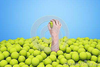 Hand appearing out of heap of yellow tennis balls holding one on blue background. Stock Photo