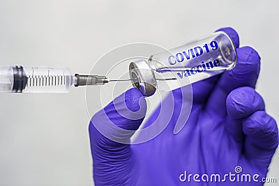 A hand in a blue medical surgical glove picks up a vaccine from an ampoule with the syringe vial inscription COVID19 vaccine Stock Photo