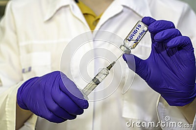 A hand in a blue medical surgical glove picks up a vaccine from an ampoule with the syringe vial inscription COVID19 vaccine Stock Photo