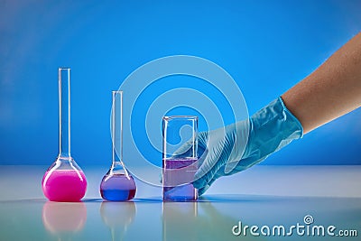 Hand in disposable glove holding beaker, purple liquid. Medical flasks, colorful chemical reagents, blue background Stock Photo