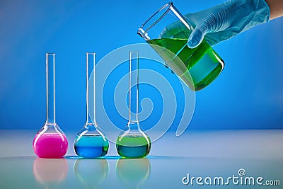Hand in disposable glove is pouring green liquid from beaker into medical flask. Colorful chemical reagents, blue Stock Photo