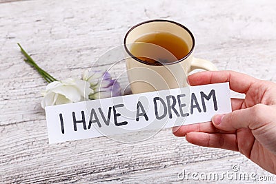 Hand holding a paper with an inscription I have a dream, against a table with a mug of black tea and flowers. Stock Photo