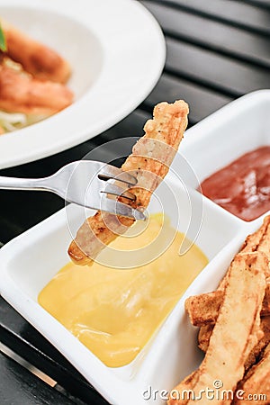 Hand holding folk french fries with in mayonnaise and ketchup sauce in the background. Mayonnaise in small round white ceramic di Stock Photo