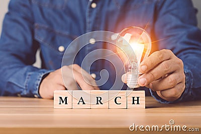 Hand holding light bulb with wooden blocks write word MARCH arranged on wooden desk with grey space wall background. Start for new Stock Photo