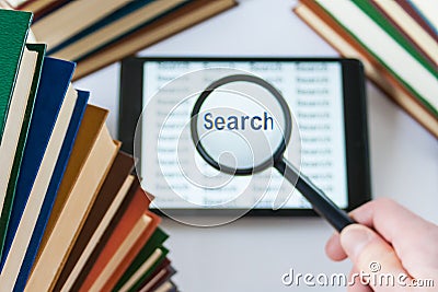 Hand holding a magnifying glass over a tablet surrounded by book Stock Photo