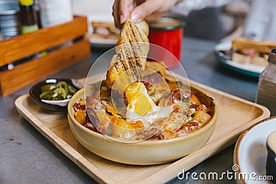 Hand holding toast dipping in Cheesy Breakfast Potatoes with Crispy Bacon and Fried egg Stock Photo