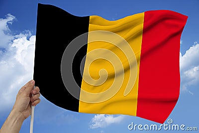 Hand holds against the background of the sky with clouds the colored flag of Belgium on the texture of the fabric, silk with waves Stock Photo