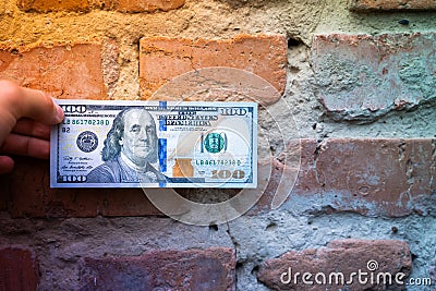 A hand holds a hundred dollar bill against the background of an old brick wall close-up. Old crumbling red brick wall and cash, us Stock Photo