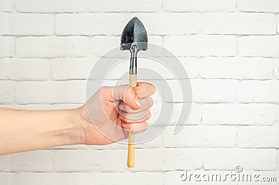 hand holds miniature gardening tool against a white brick wall. shovel in a woman's hand Stock Photo