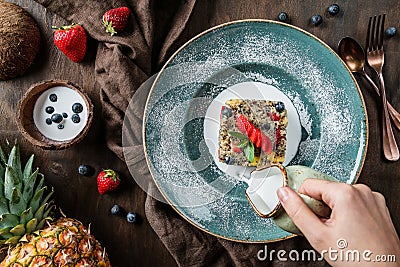 Hand pouring a coconut milk into quinoa porridge with fruits, strawberries, blueberry, pineapple over brown wooden background Stock Photo